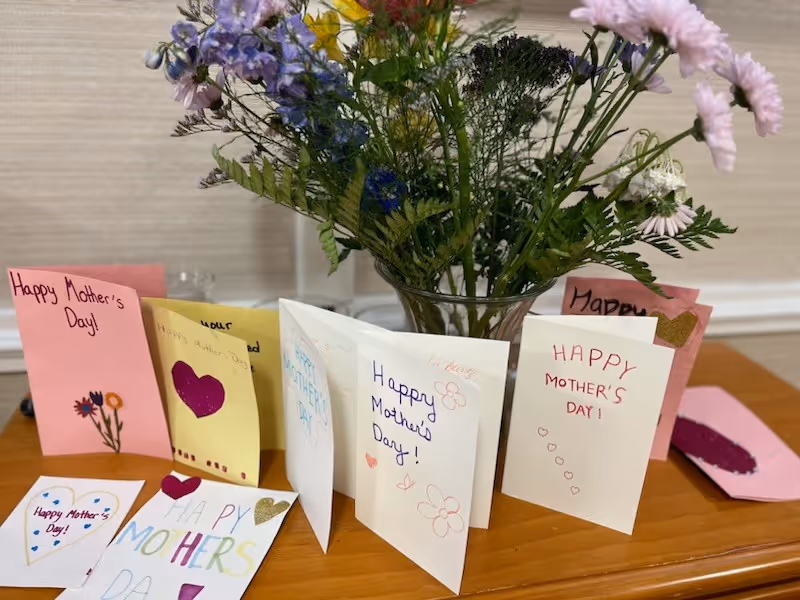 A wooden table displaying a variety of handmade Mother's Day cards with colorful drawings and messages, arranged in front of a vase filled with a mixed bouquet of flowers including pink, purple, yellow, and white blooms.