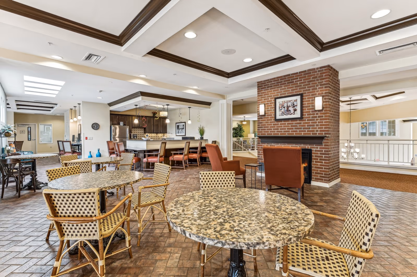 A spacious common area in a senior living facility featuring round tables with woven chairs, a brick fireplace with two armchairs, and a kitchen area with bar stools and stainless steel appliances. The room has a warm, inviting atmosphere with recessed lighting and decorative ceiling beams.
