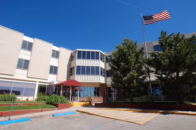Exterior view of a multi-story building with large windows and an entrance covered by a red awning. There are trees and an American flag on a flagpole in front of the building under a clear blue sky.