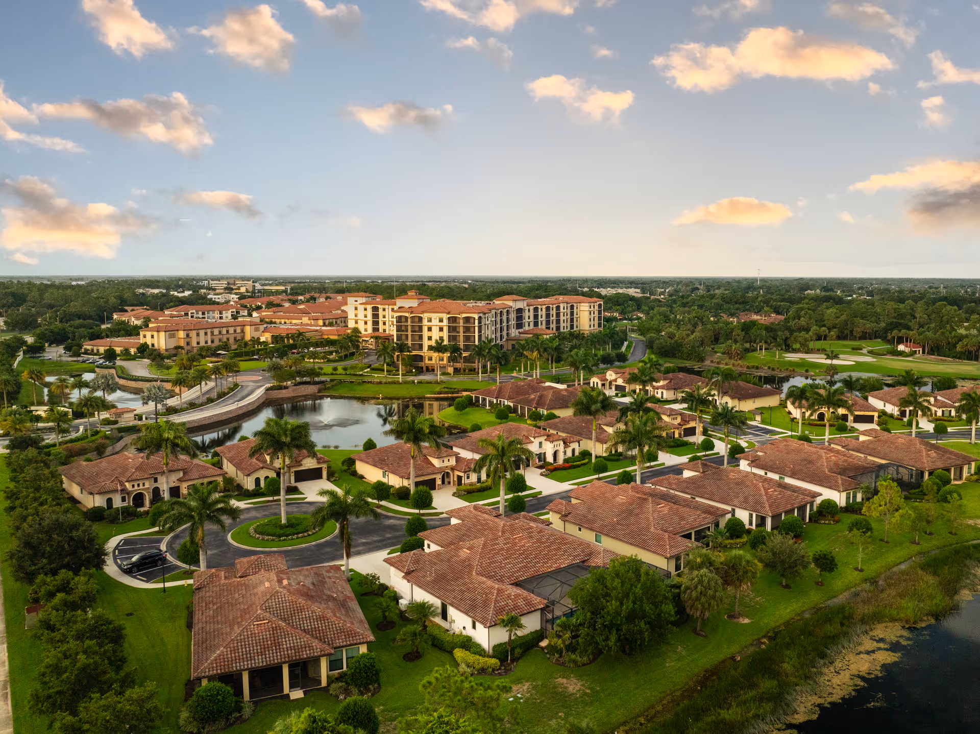 Aerial view of The Arlington of Naples campus with Mediterranean-style villas, palm trees, a central pond, and a multi-story building in the background.