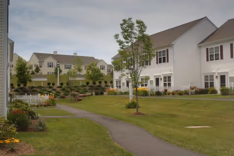 A landscaped outdoor area of a senior living facility with paved walking paths, green lawns, small trees, and white multi-unit residential buildings with red shutters in the background under a cloudy sky.