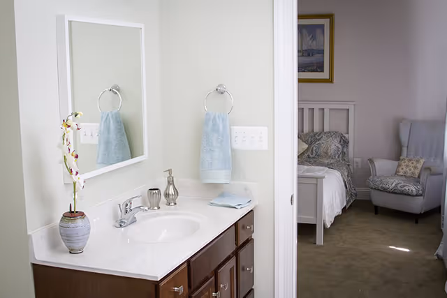 Bathroom vanity with sink, mirror, towel and vase, opening into a bedroom with a bed and armchair.
