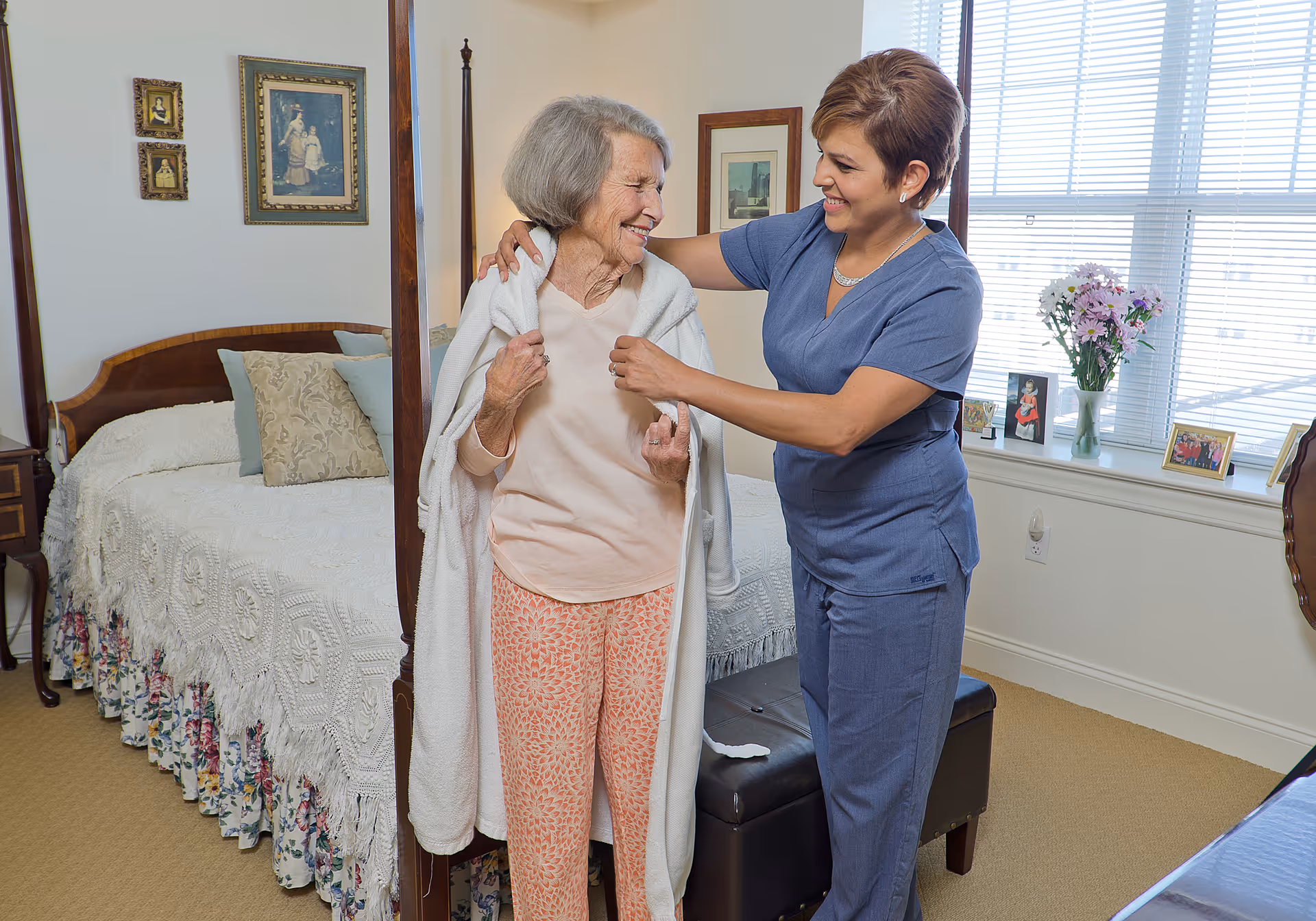 An elderly woman standing in a bedroom with a caregiver who is helping her put on a white robe. The bedroom has a bed with a white crocheted bedspread and floral bed skirt, framed pictures on the wall, and a window with blinds and a windowsill decorated with flowers and framed photos.