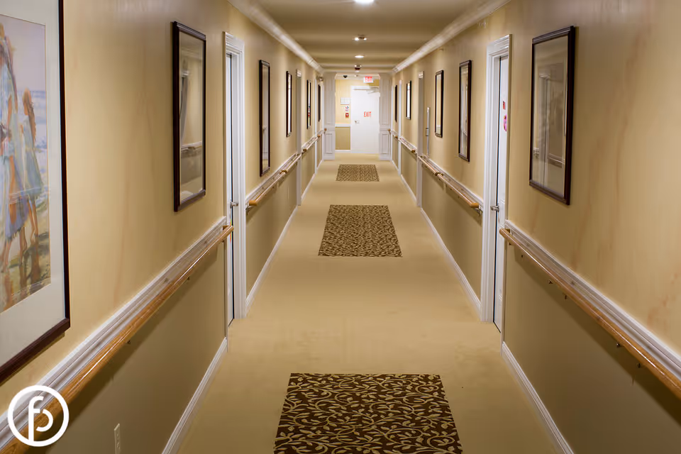 A long, well-lit hallway in a senior living facility with beige walls and carpeted floor. The hallway has handrails on both sides and framed artwork hanging on the walls. Several white doors line the corridor, and an exit sign is visible at the far end.