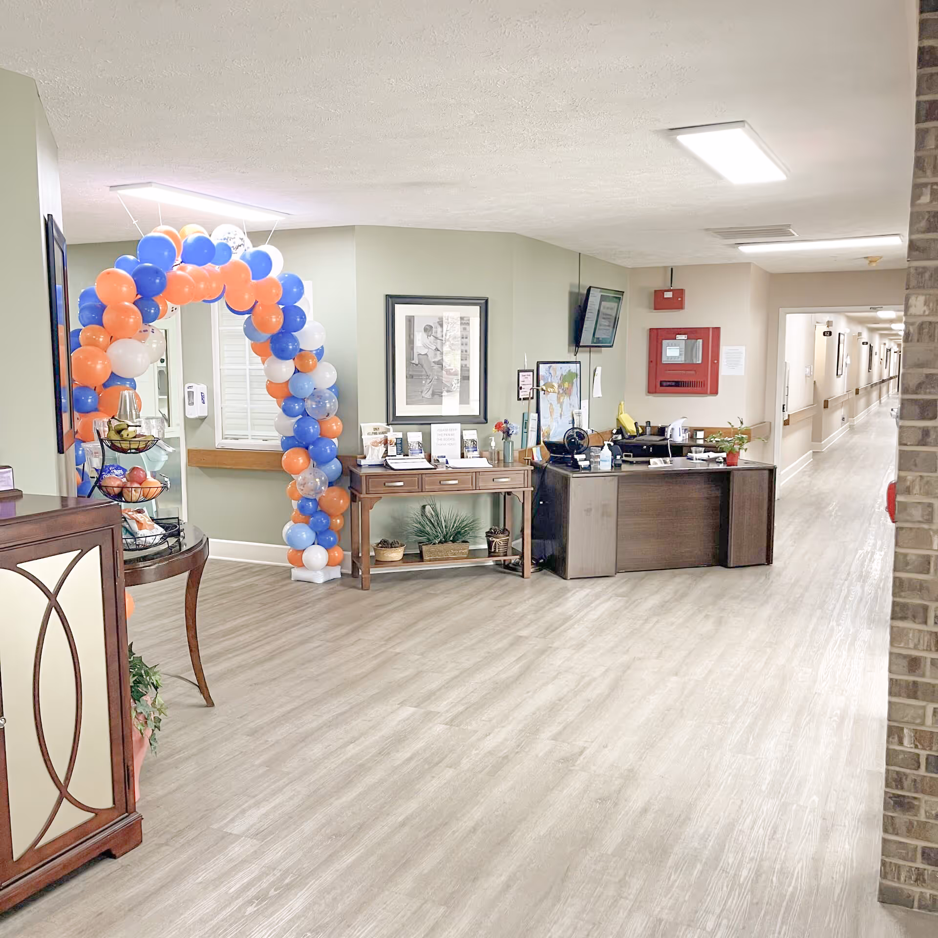 Interior hallway of a senior living facility with light wood flooring, a reception desk, a table with informational brochures, a colorful balloon arch in blue, orange, and white, and a long corridor with doors on the right side.