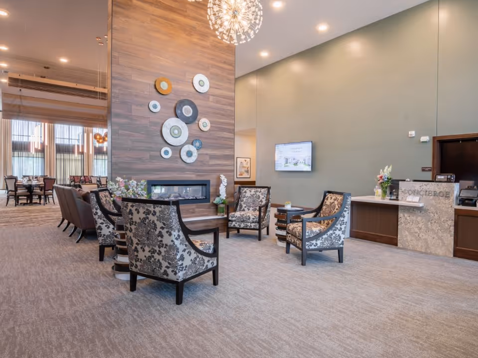 Bright senior living lobby with patterned armchairs arranged around a fireplace and a concierge desk on the right.