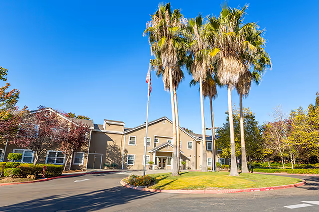 Exterior view of a senior living facility building with beige siding, multiple windows, and a covered entrance. There are tall palm trees and an American flag on a flagpole in front of the building, with a circular driveway and landscaped greenery under a clear blue sky.