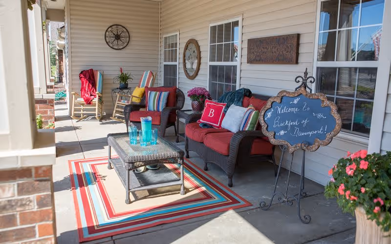 A cozy outdoor patio area with wicker furniture including a loveseat and chairs with red cushions and colorful striped pillows. A small table with blue glassware sits on a striped outdoor rug. There are decorative plants and flowers, a wall clock, and a chalkboard sign that reads 'Welcome to Backyard of Davenport!'