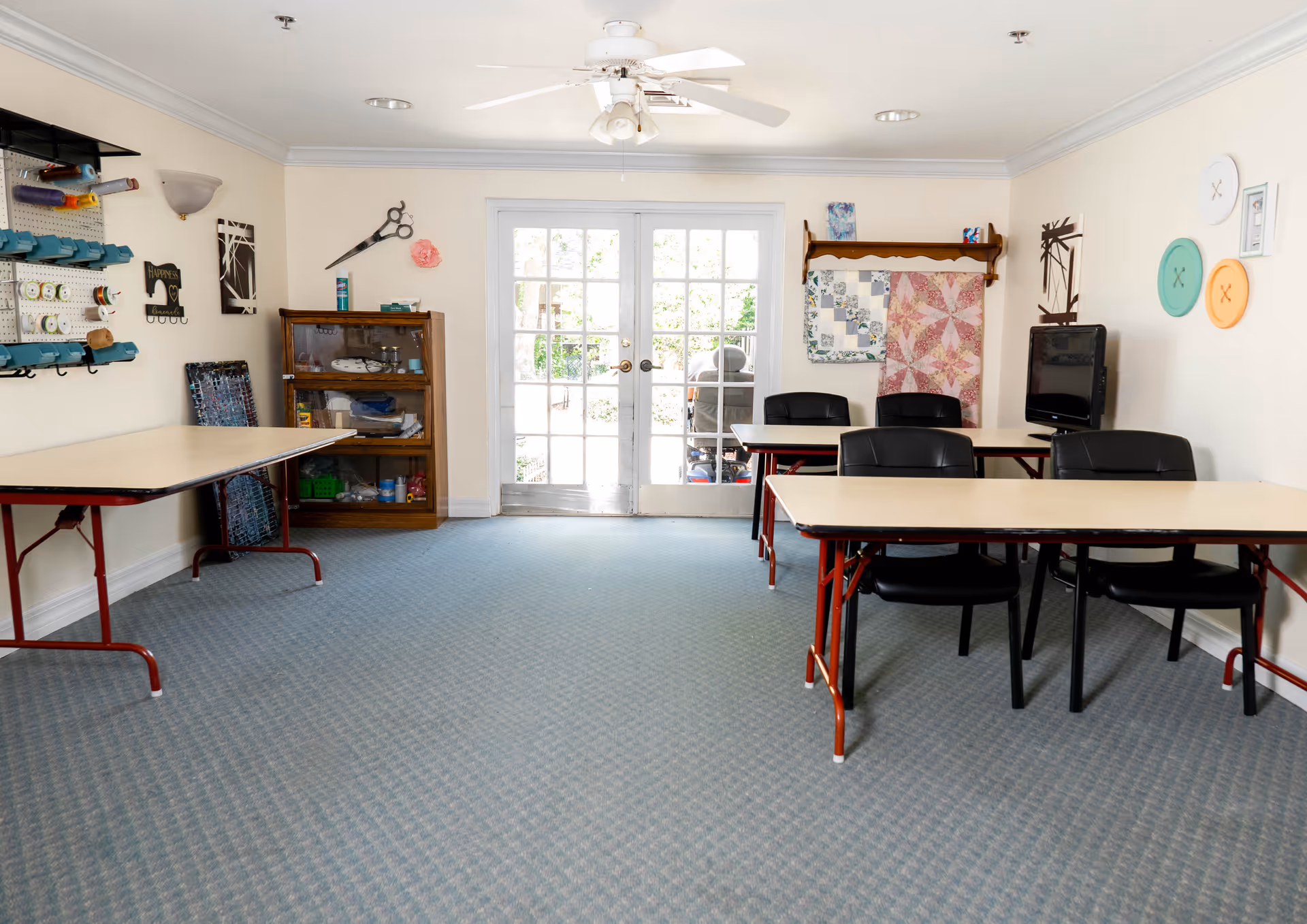 A craft room with tables and chairs arranged for group activities. The room has a blue carpet, white walls, and a ceiling fan. On the walls are various sewing and craft supplies, including large decorative scissors, quilted wall hangings, and oversized button decorations. French doors at the back let in natural light and provide a view of an outdoor area.