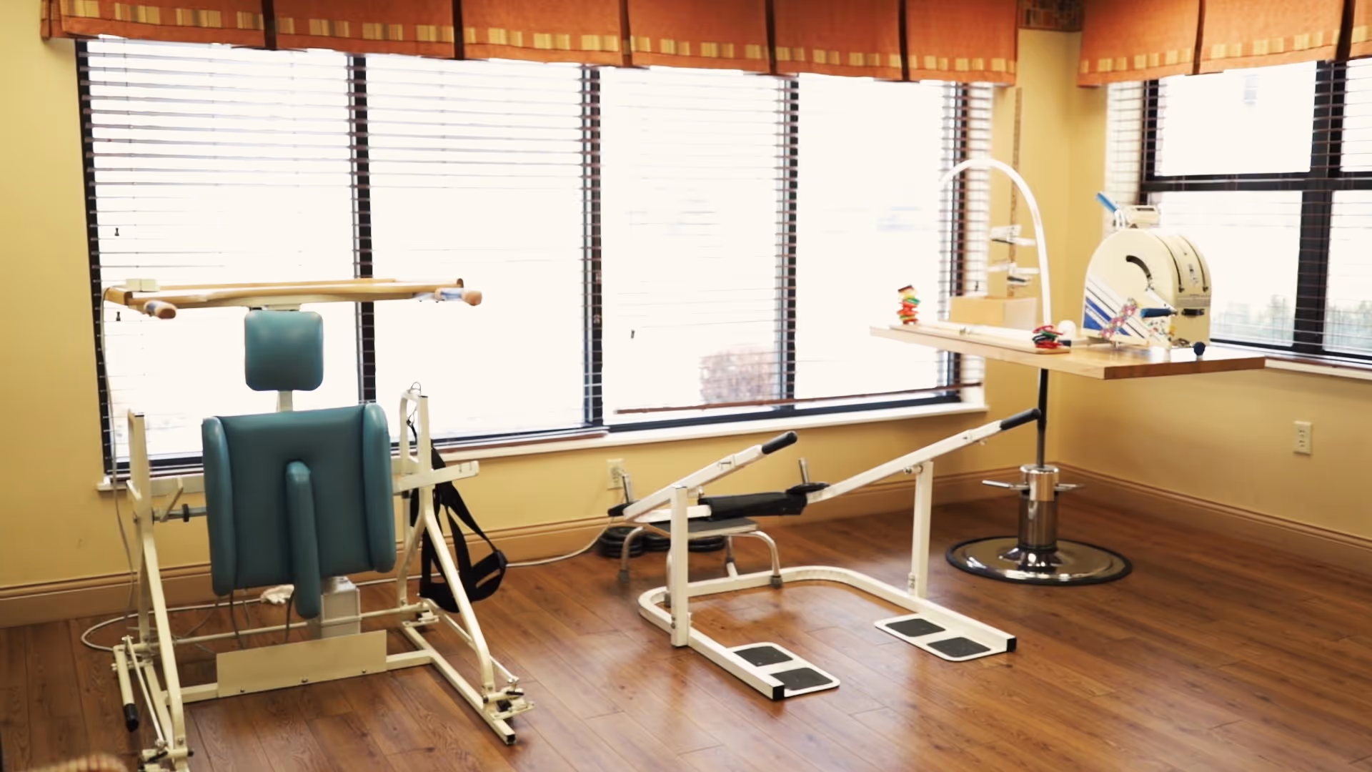 A bright room with large windows covered by blinds and valances, featuring physical therapy equipment including a padded exercise chair with wooden bars and a leg press machine, along with a table holding therapy tools and devices.