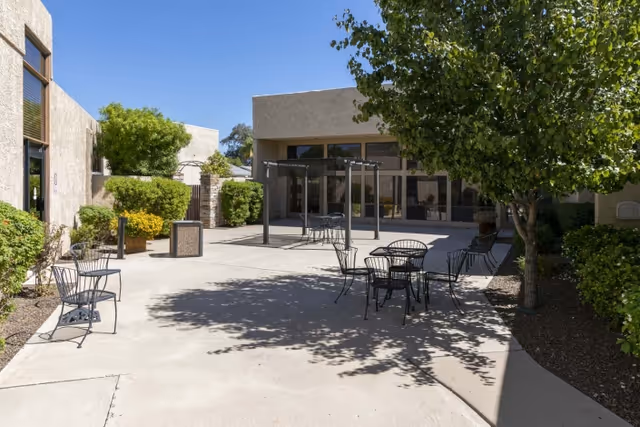 Outdoor courtyard with metal tables and chairs, trees, and building entrances under a clear sky.