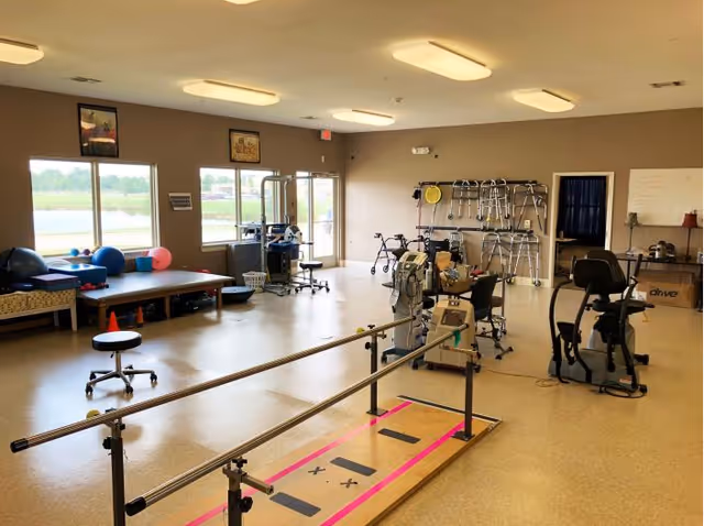 A spacious rehabilitation or physical therapy room with exercise equipment including parallel bars, stationary bikes, walkers, therapy balls, and padded tables. The room has large windows letting in natural light and a beige floor and walls.