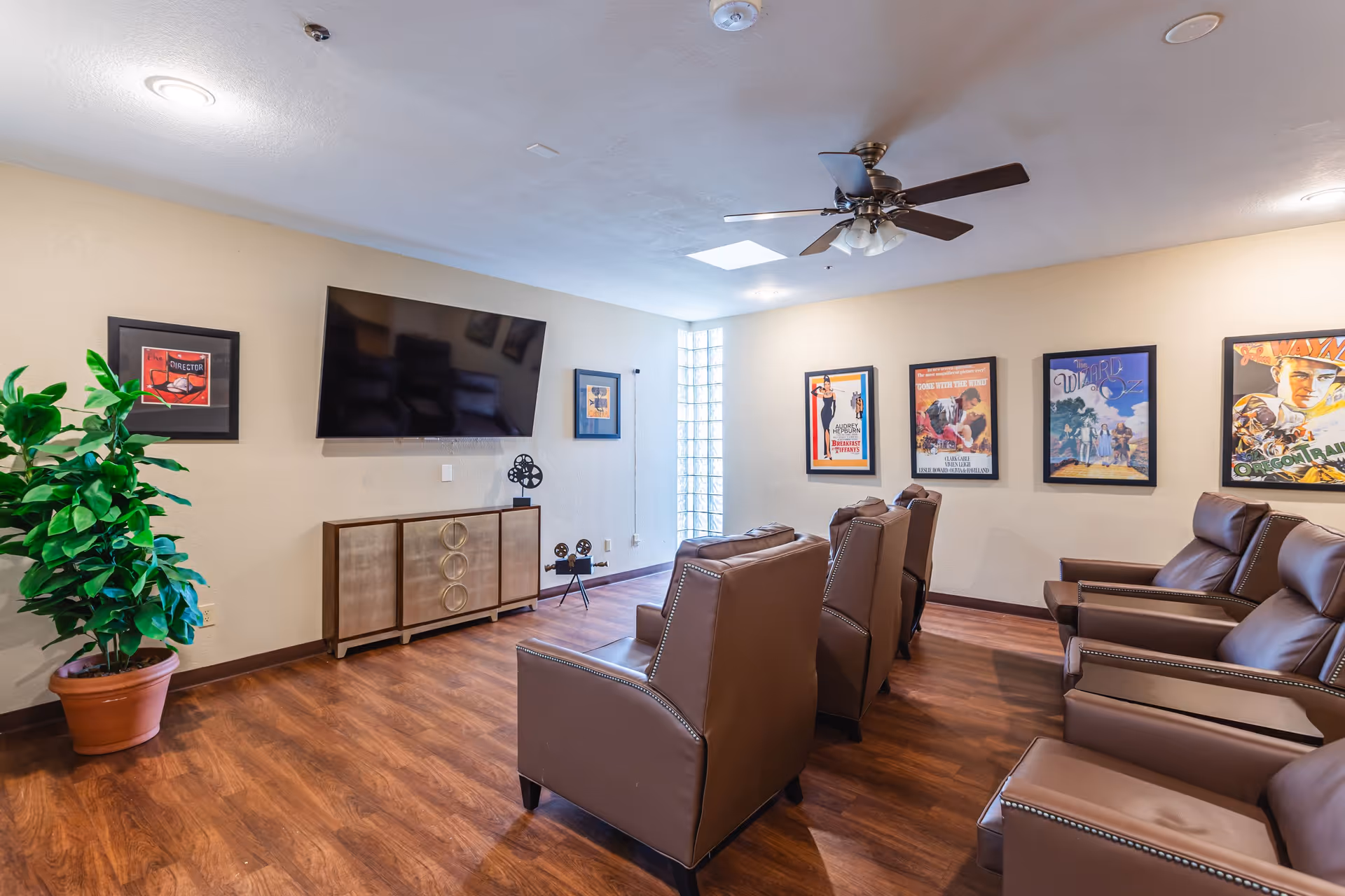 A cozy media room with several brown leather recliner chairs arranged in rows facing a large flat-screen TV mounted on a beige wall. The room has wood flooring, a ceiling fan with lights, framed movie posters on the walls, a potted plant in the corner, and decorative film reel sculptures on a cabinet beneath the TV.