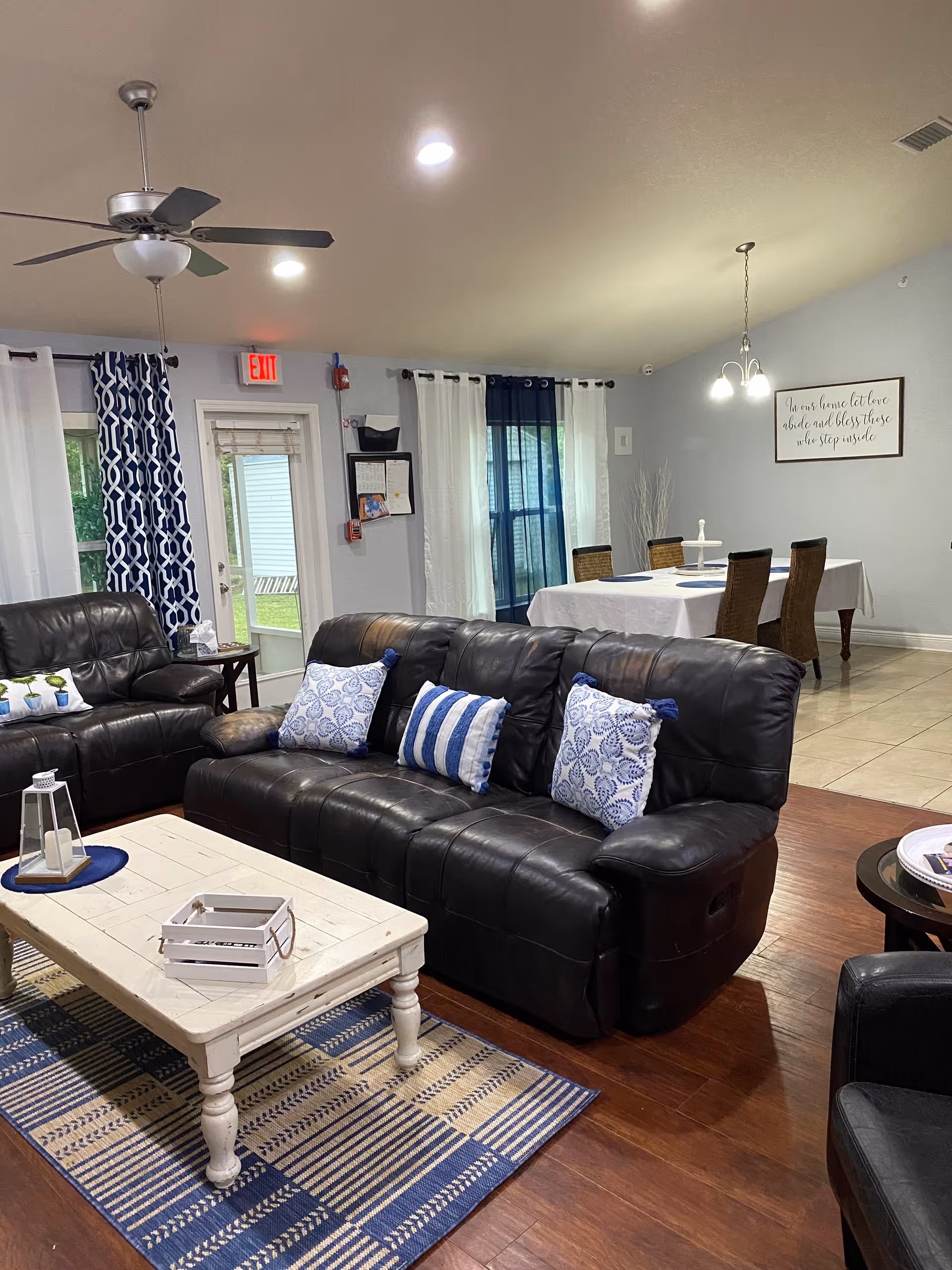 Interior view of a living room and dining area in Mariner Palms Assisted Living Facility. The living room features a black leather sofa and loveseat with blue and white patterned pillows, a white wooden coffee table on a blue and beige patterned rug, and a ceiling fan with lights. The dining area has a table covered with a white tablecloth and four wicker chairs. The walls are light gray with white and blue curtains on the windows and a framed sign on the wall. There is a door with an exit sign above it leading outside.
