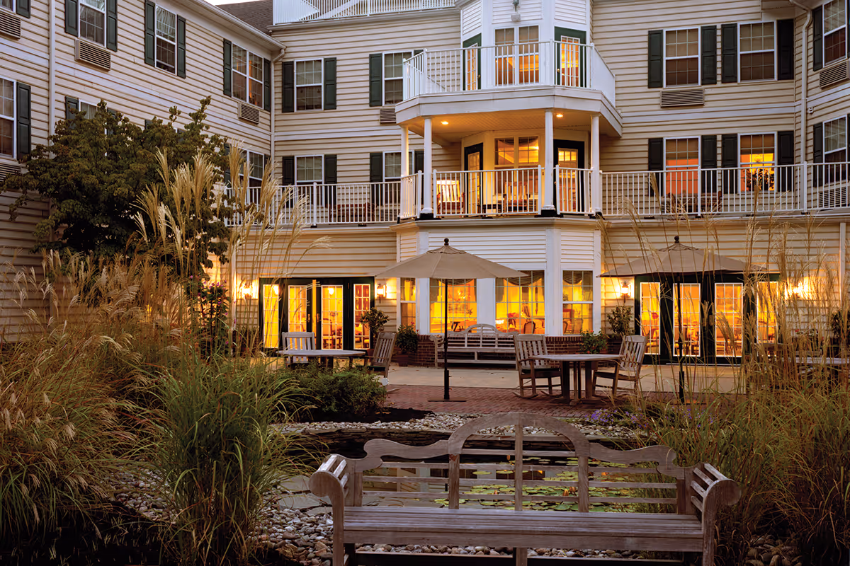 Outdoor courtyard area of a senior living facility with wooden benches, tables with umbrellas, tall grasses, and a small pond in the foreground. The building in the background has multiple windows with warm lights glowing inside and balconies with white railings.