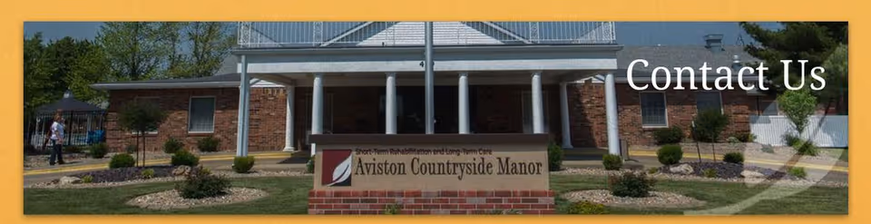 Front exterior view of a brick building with white columns and a sign in front that reads 'Aviston Countryside Manor'. There is a person walking on the left side near a tent and some landscaping with bushes and grass.
