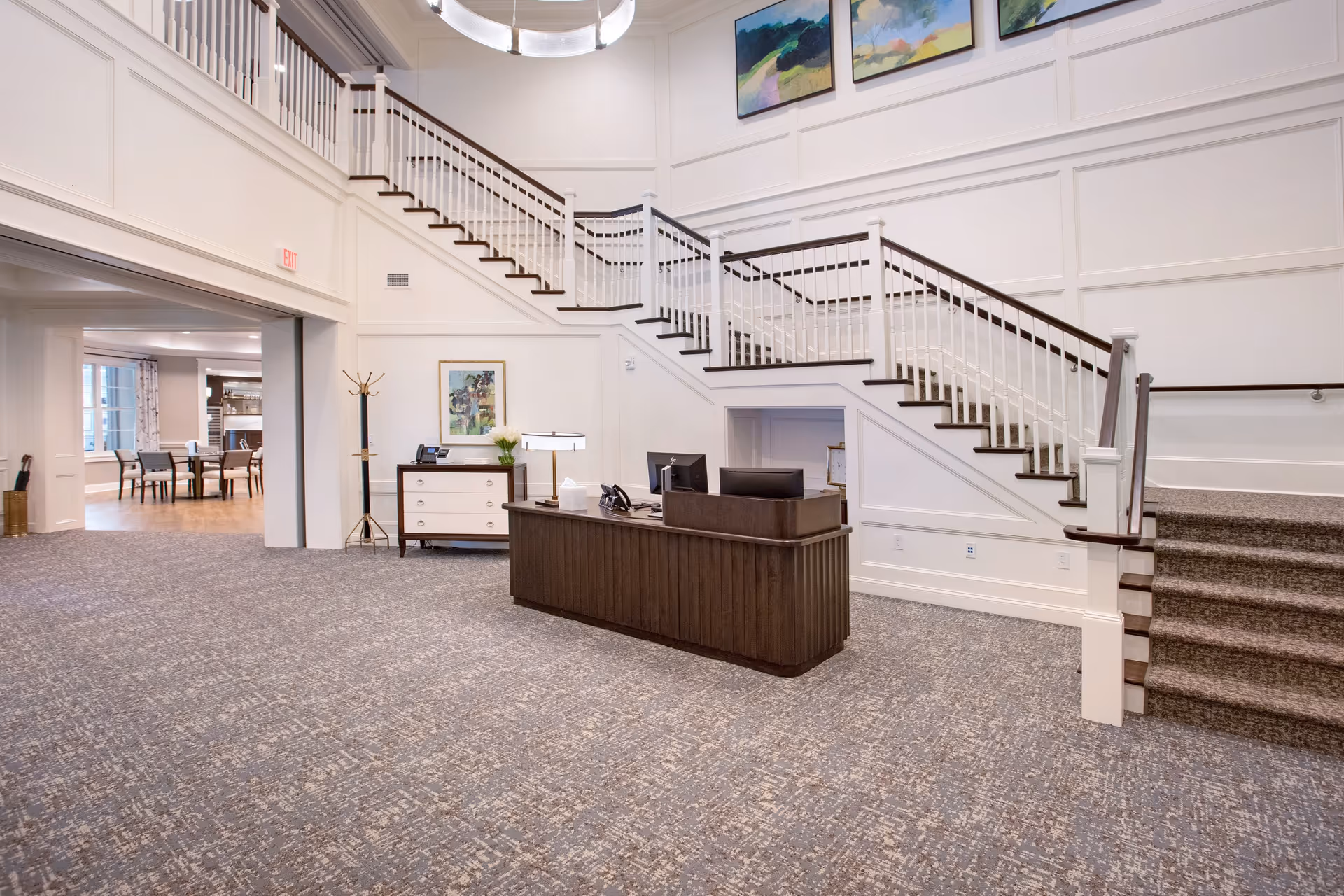 Interior view of a senior living facility lobby with a dark wooden reception desk in the center, a staircase with white railings and dark wooden handrails leading to an upper floor, and a small chest of drawers with a lamp and flowers against the wall. The area has carpeted flooring and light-colored walls with decorative paneling and paintings above the staircase. In the background, there is a dining area with tables and chairs.