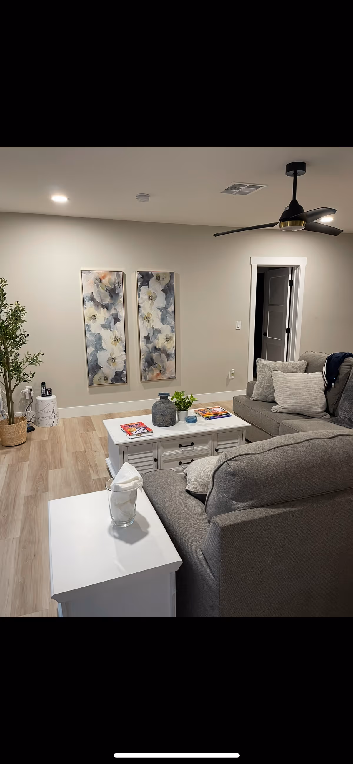 Modern living room with gray sofas, white coffee and side tables, floral wall art, potted plant, and a ceiling fan.