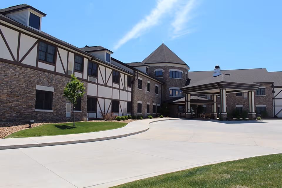 Front entrance and driveway of a multi-story brick and Tudor-style senior living building under a blue sky.