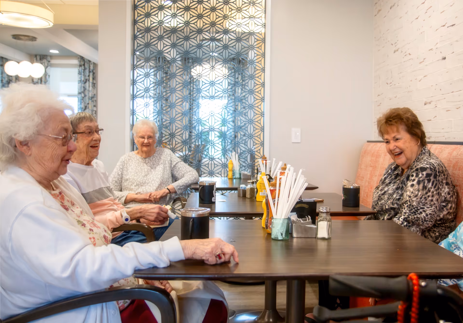 Four elderly women sitting around a dining table in a well-lit room with patterned curtains and a decorative partition in the background. They appear to be engaged in conversation and enjoying each other's company. The table has condiments, straws, and cups on it.