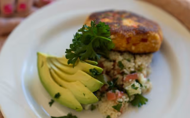 A close-up of a plated meal featuring sliced avocado, a serving of couscous salad with chopped tomatoes and parsley, and a golden-brown patty garnished with a sprig of parsley.