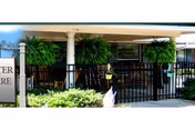 Front porch area of Goodwater Healthcare Center featuring a covered entrance with white columns, hanging green ferns, black metal railing, and a sign partially visible on the left side.