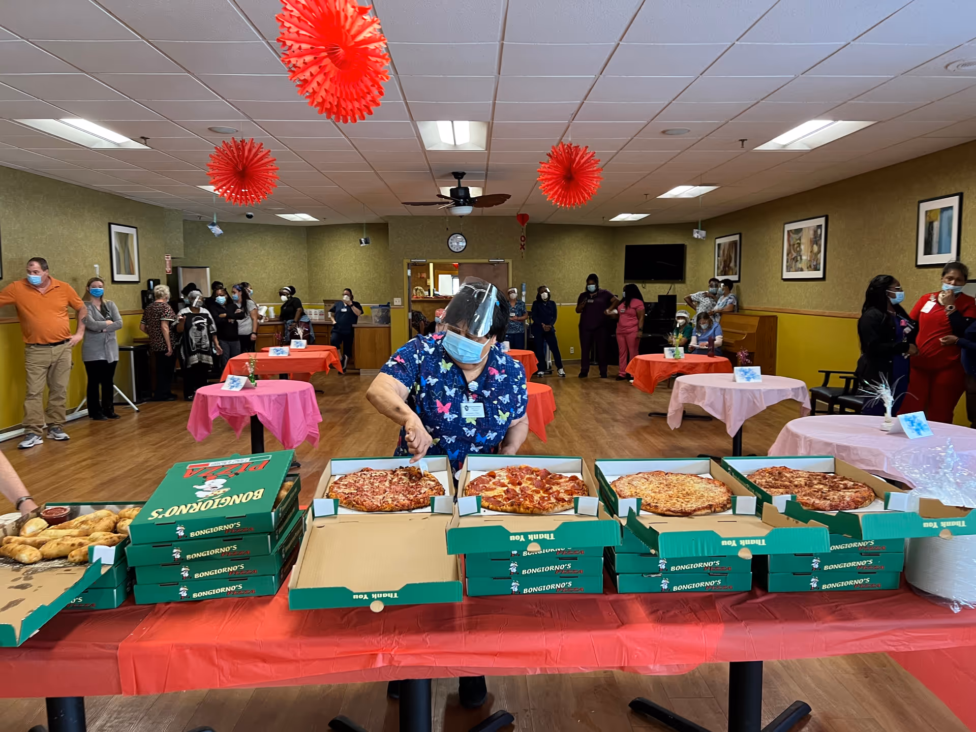A dining area decorated with red hanging paper decorations and tables covered with pink and red tablecloths. A woman wearing a face shield and mask is cutting pizza from several open pizza boxes on a table in the foreground. People wearing masks are standing and sitting around the room, some near the walls and others at tables.