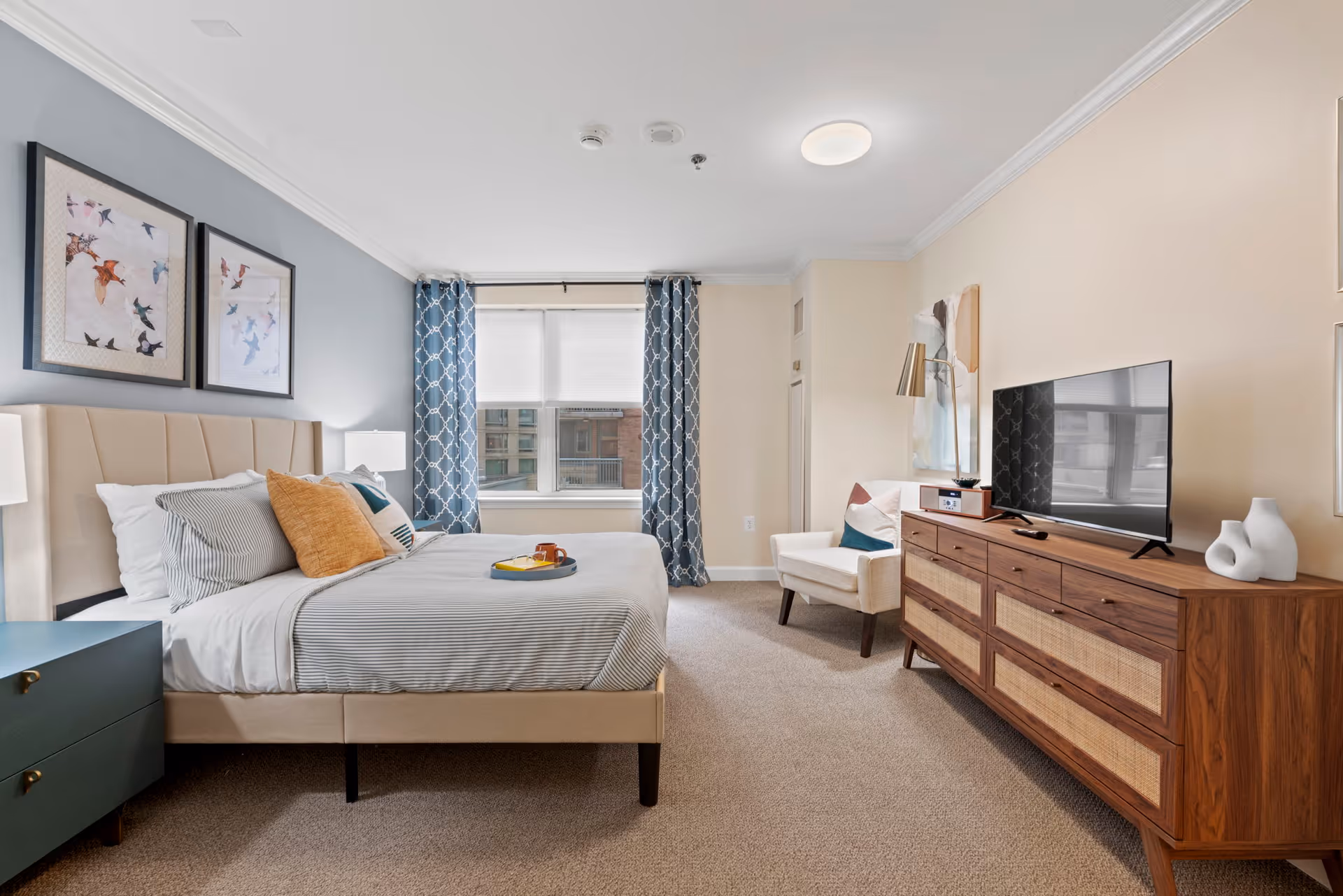 A neatly arranged bedroom with a beige upholstered bed featuring striped bedding and multiple pillows. There are two framed bird artworks on the blue accent wall above the bed. A dark green nightstand with a lamp is beside the bed. Across from the bed is a wooden dresser with a flat-screen TV and decorative items on top. A white armchair with a colorful pillow is placed near the dresser. The room has a window with blue patterned curtains and a beige carpeted floor.