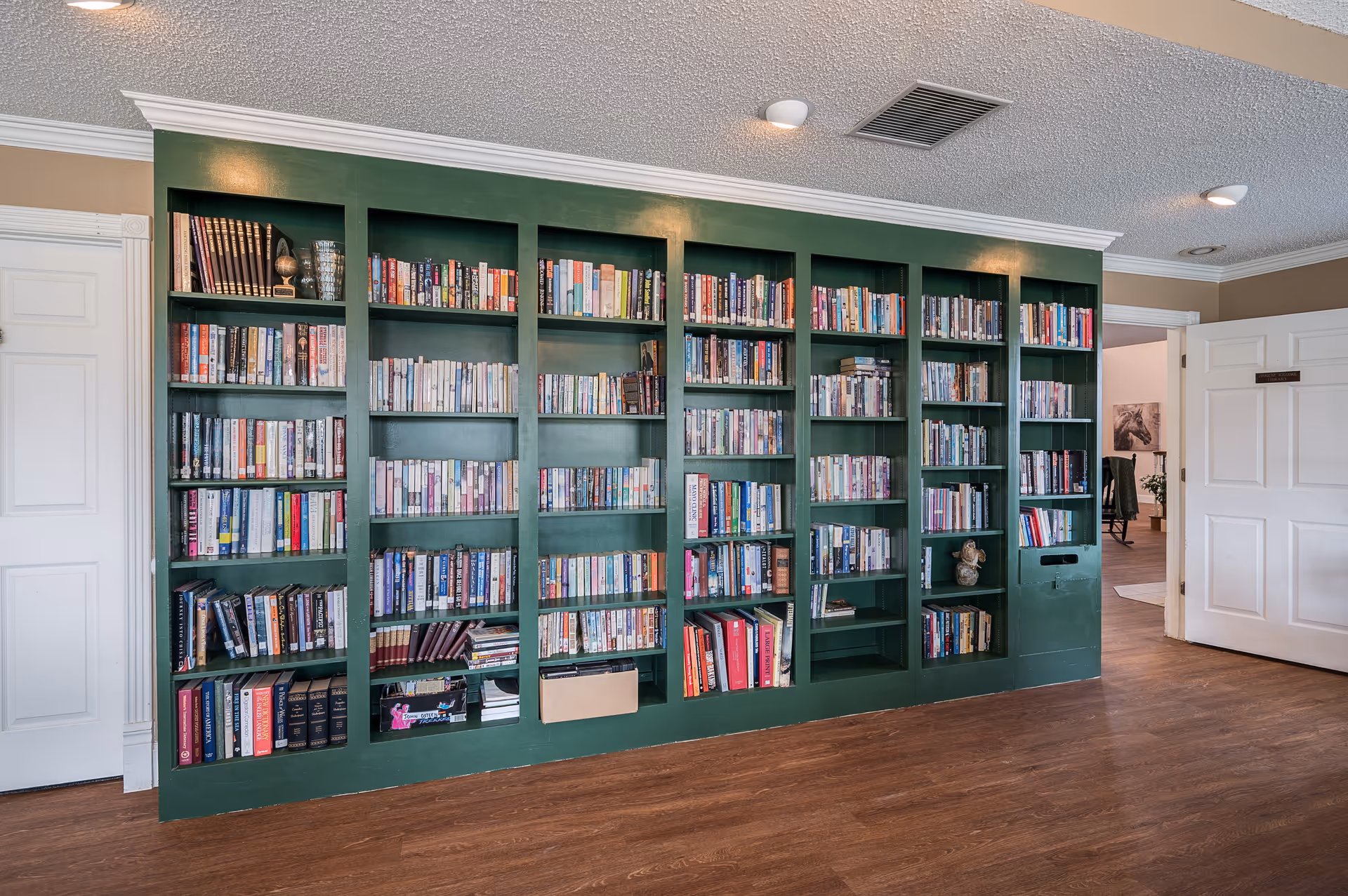 Interior view of a room with a large green built-in bookshelf filled with books. The room has wooden flooring, beige walls, white trim, and two white doors, one of which is open showing another room with a chair and artwork on the wall.