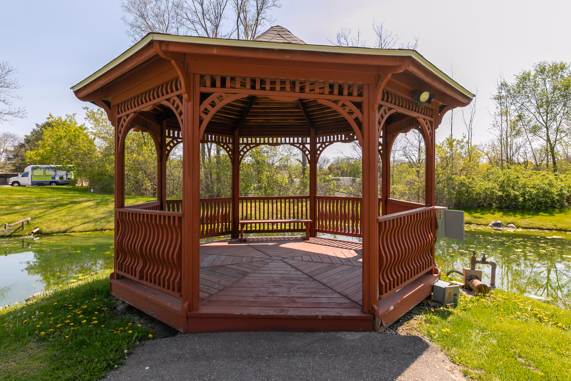 A red wooden gazebo with decorative railings and a bench inside, situated next to a small pond surrounded by green grass and trees under a clear sky.