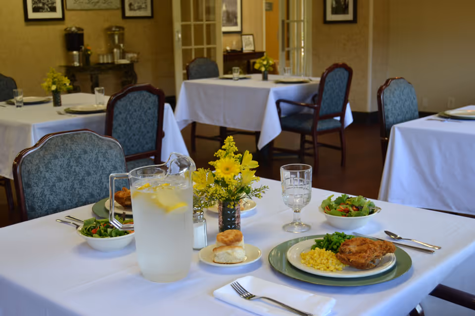 A dining room with tables covered in white tablecloths, set with plates of food including fried chicken, corn, green beans, salad, and biscuits. There is a pitcher of water with lemon slices, glasses, silverware, and a small vase with yellow flowers on the table. The room has upholstered chairs and framed pictures on the walls.