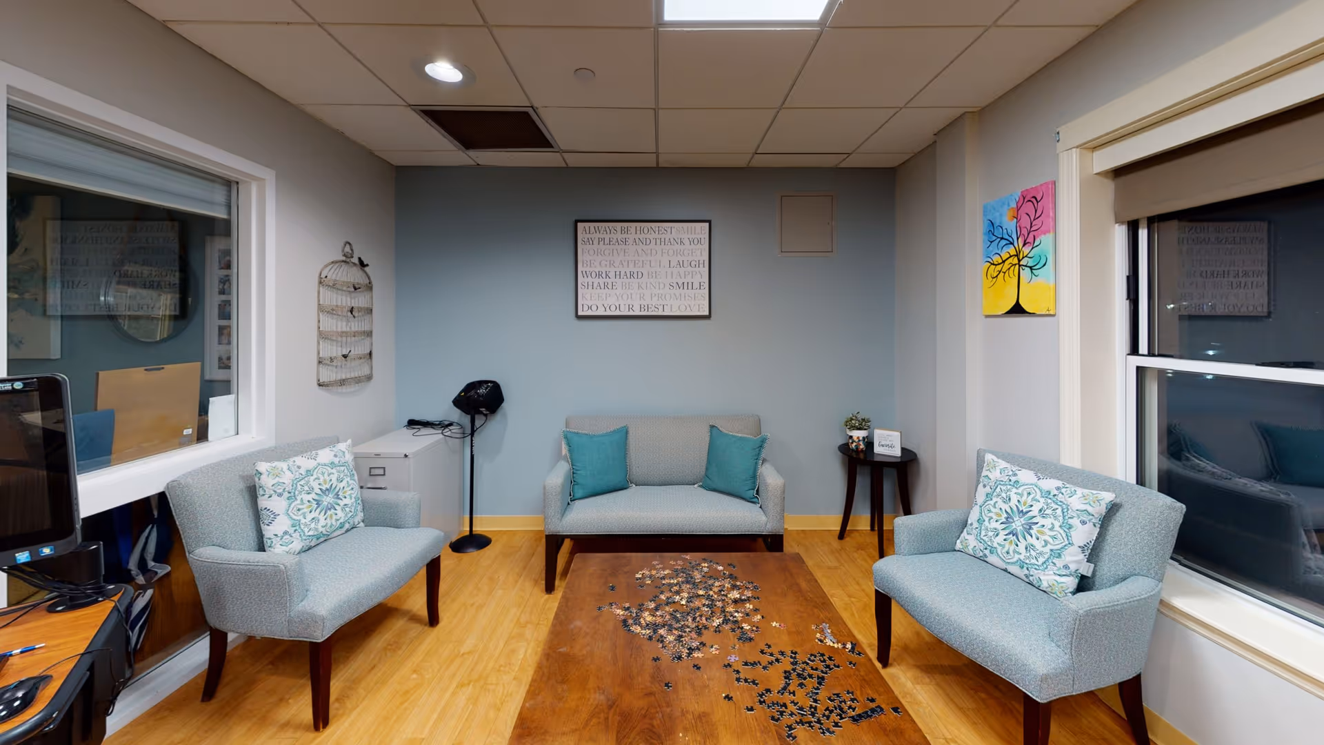 A small, cozy living room with three light blue upholstered chairs arranged around a wooden coffee table with a partially completed jigsaw puzzle on it. The room has light wood flooring, a blue accent wall with a framed inspirational quote, and two windows with white trim. There is a small round side table with a plant and a sign, a colorful painting of a tree on the right wall, and a computer workstation visible through a window on the left.