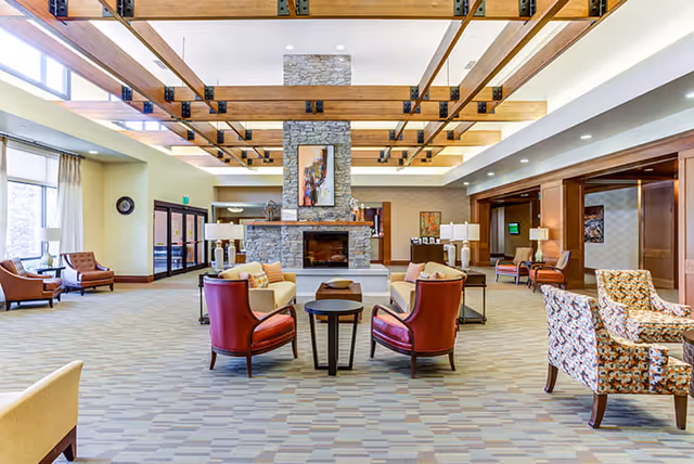Spacious senior living facility common area with a large stone fireplace in the center, surrounded by comfortable seating including armchairs and sofas. The room features wooden ceiling beams, large windows letting in natural light, and a patterned carpet floor.