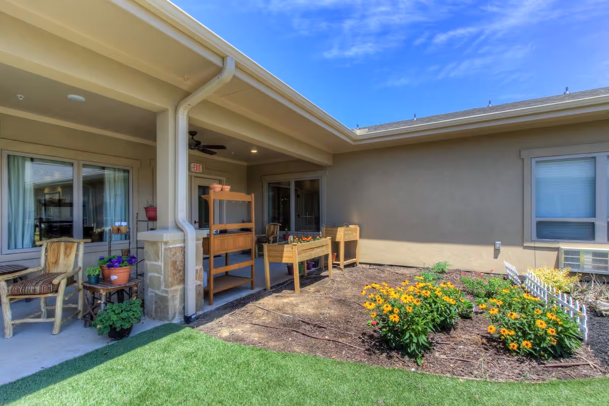 Covered patio and outdoor courtyard with seating, potted plants, raised planters, and flower beds at an assisted living facility.