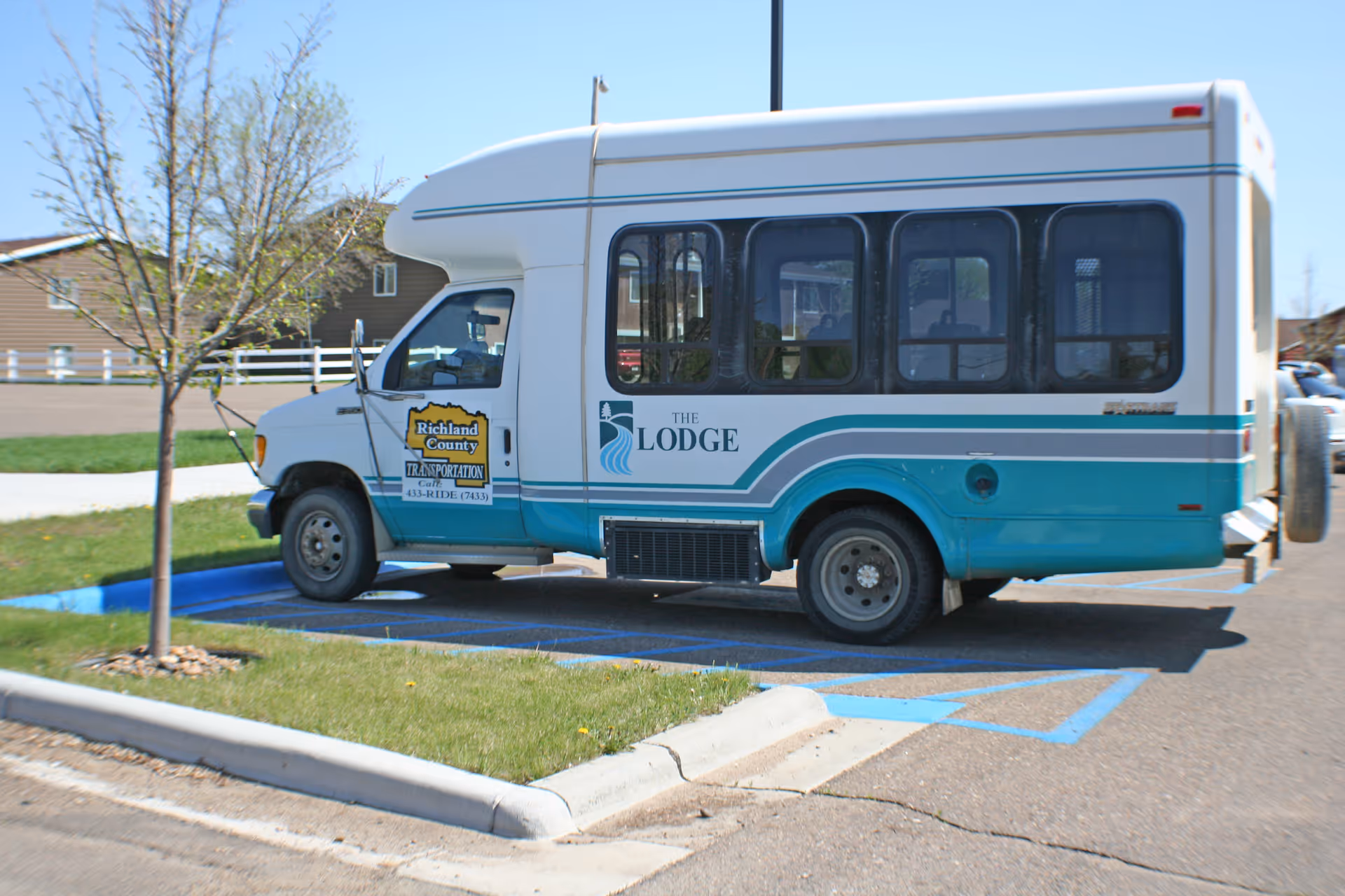 A teal-and-white shuttle van labeled "The Lodge" parked in an outdoor handicapped parking space by a small tree.