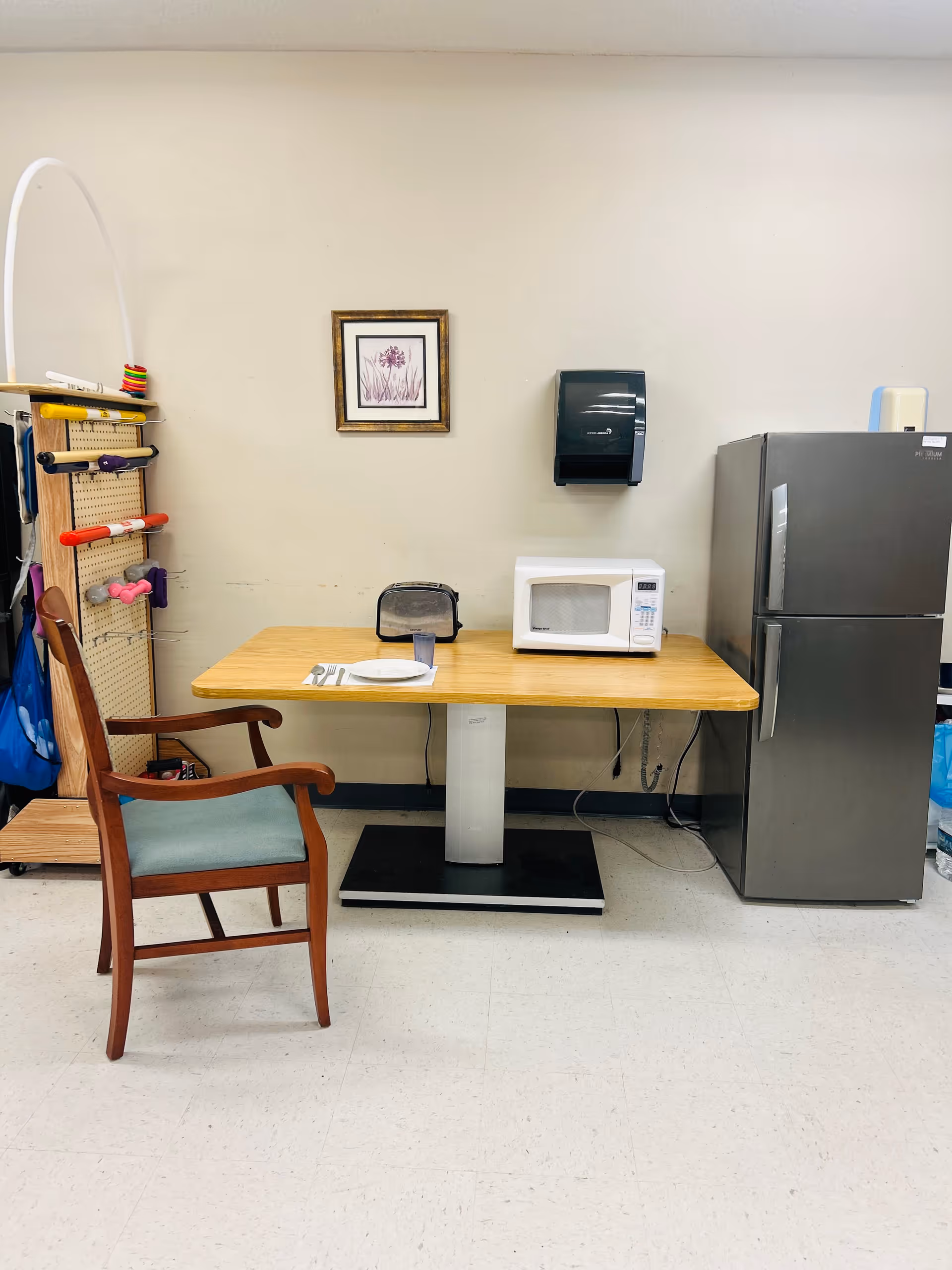 A small dining area with a wooden table set against a beige wall. On the table are a white microwave, a black toaster, a plate with utensils, and a glass. To the left of the table is a wooden chair with green upholstery. On the wall above the table is a framed picture of purple flowers and a black paper towel dispenser. To the right of the table is a stainless steel refrigerator. On the left side of the image, there is a rack holding various exercise equipment such as dumbbells and resistance bands.