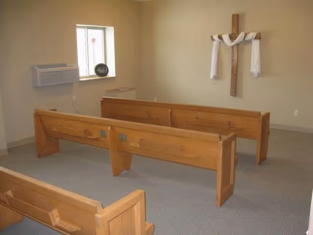 Small chapel room with wooden pews arranged in rows facing a wooden cross draped with white cloth on the wall. There is a small window with an air conditioning unit below it and a decorative plate on the windowsill.