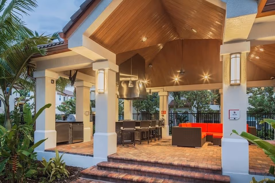 Covered outdoor pavilion with a wooden vaulted ceiling featuring a bar and grill area, high stools and a red sectional sofa surrounded by tropical plants.