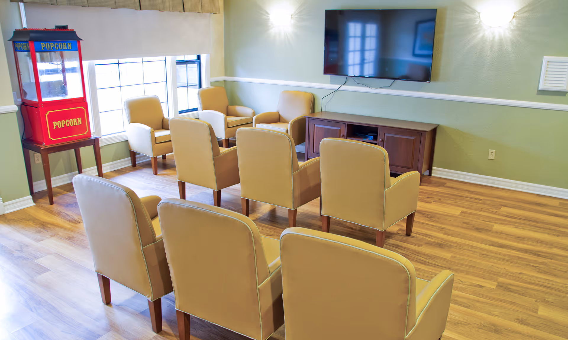 A small lounge area with eight beige armchairs arranged in two rows facing a wall-mounted flat screen TV. There is a wooden cabinet below the TV and a red popcorn machine on a small table near the window. The room has light green walls, wooden flooring, and two wall-mounted light fixtures.