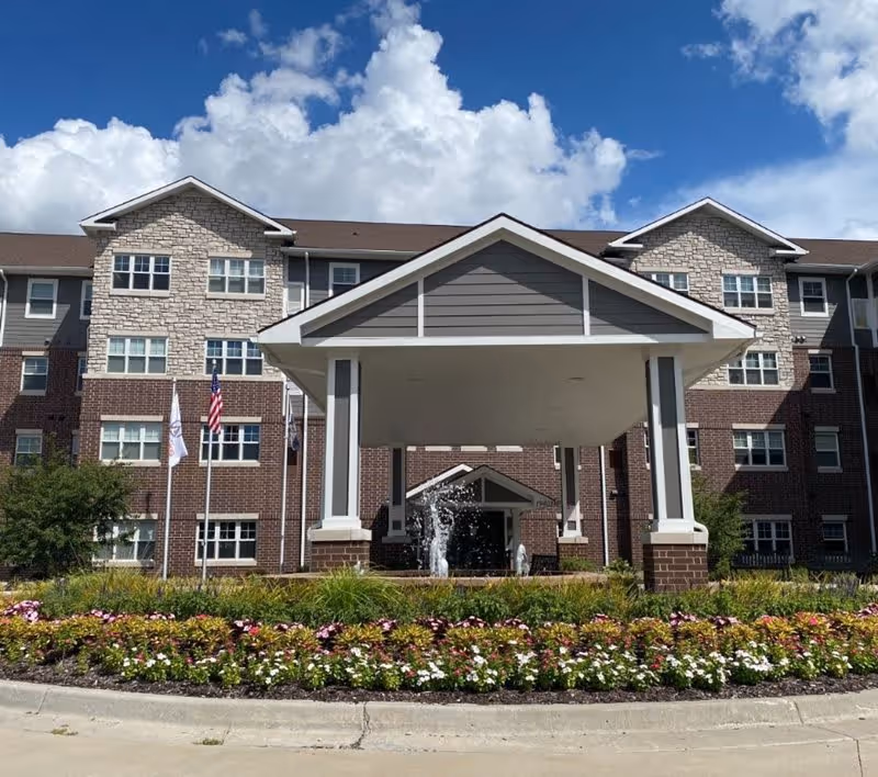 Front exterior view of a multi-story senior living facility building with a covered entrance, a water fountain, and a landscaped flower bed in front. The building has a combination of brick and stone facade under a partly cloudy blue sky.