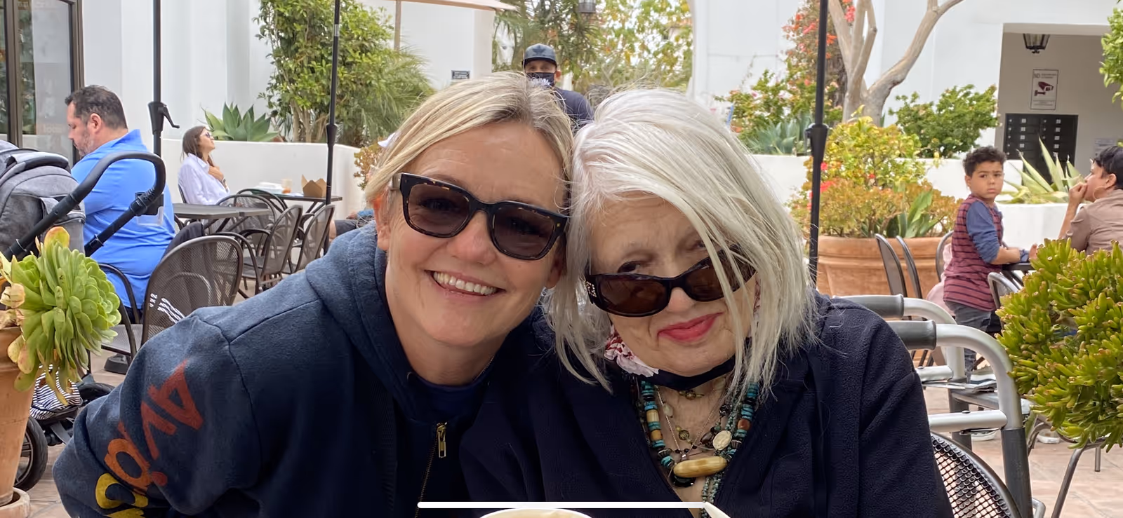 Two women wearing sunglasses sitting closely together and smiling at an outdoor cafe with tables, chairs, plants, and other people in the background.