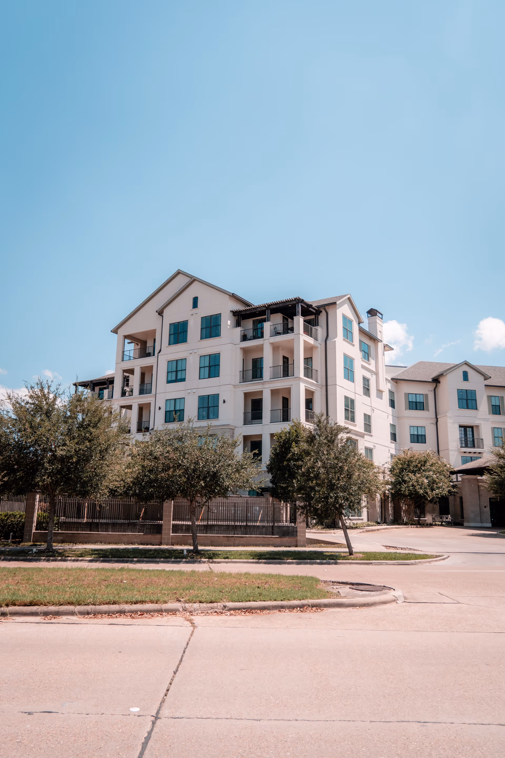 Front exterior of a multi-story senior living building with balconies, trees, and a clear blue sky.
