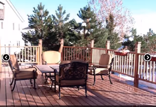 Outdoor wooden deck with a table and four cushioned chairs surrounded by a wooden railing, with trees and part of a building visible in the background.