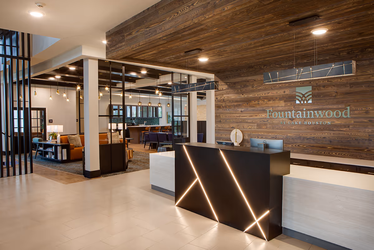 Reception area of Fountainwood at Lake Houston featuring a modern black front desk with diagonal light strips, a wooden accent wall with the facility's name and logo, and a seating area with orange couches and pendant lights in the background.