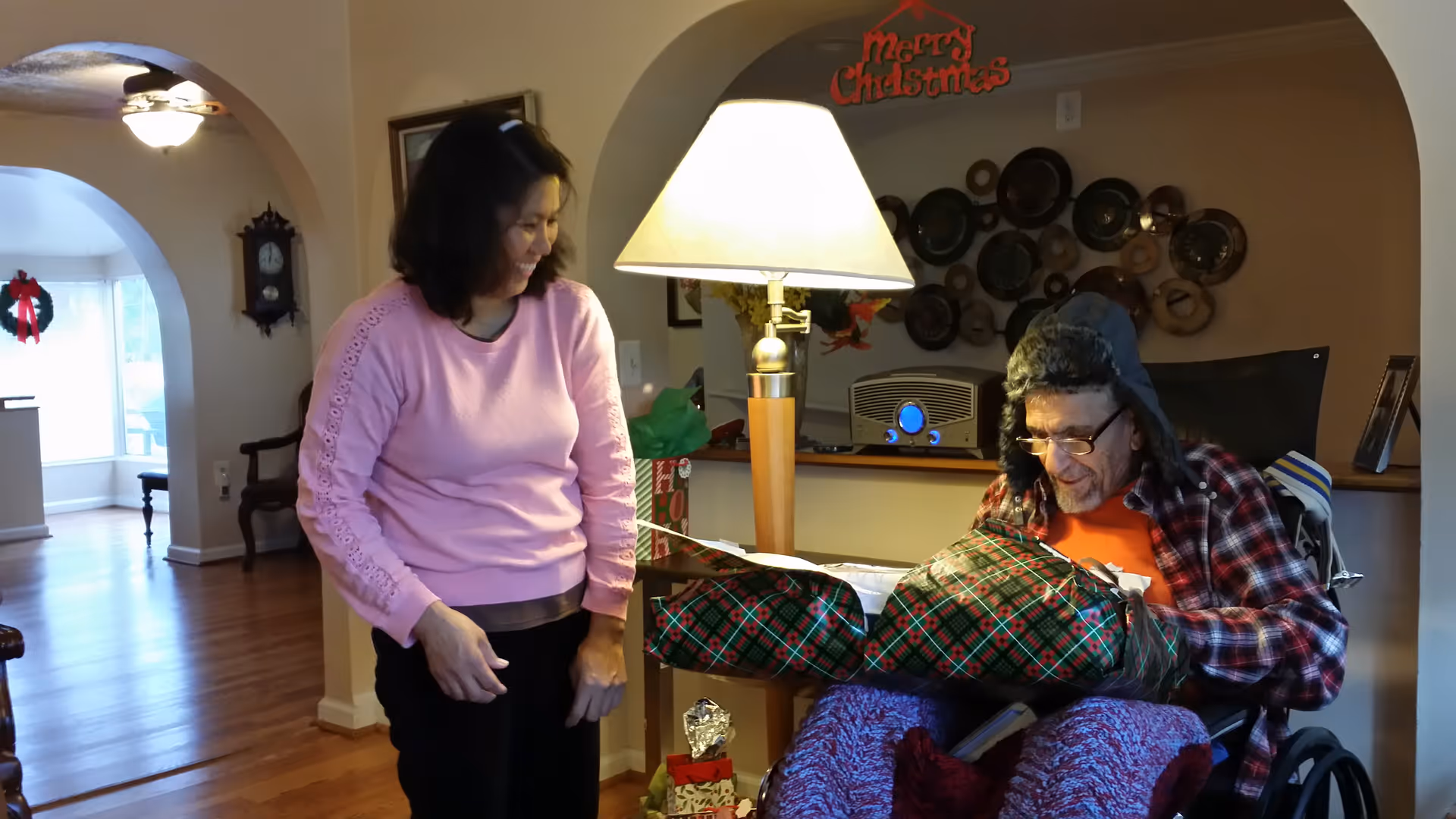 A woman in a pink sweater smiling and standing next to an elderly man in a wheelchair who is opening a Christmas gift wrapped in green and red plaid paper inside a warmly lit living room decorated for Christmas.