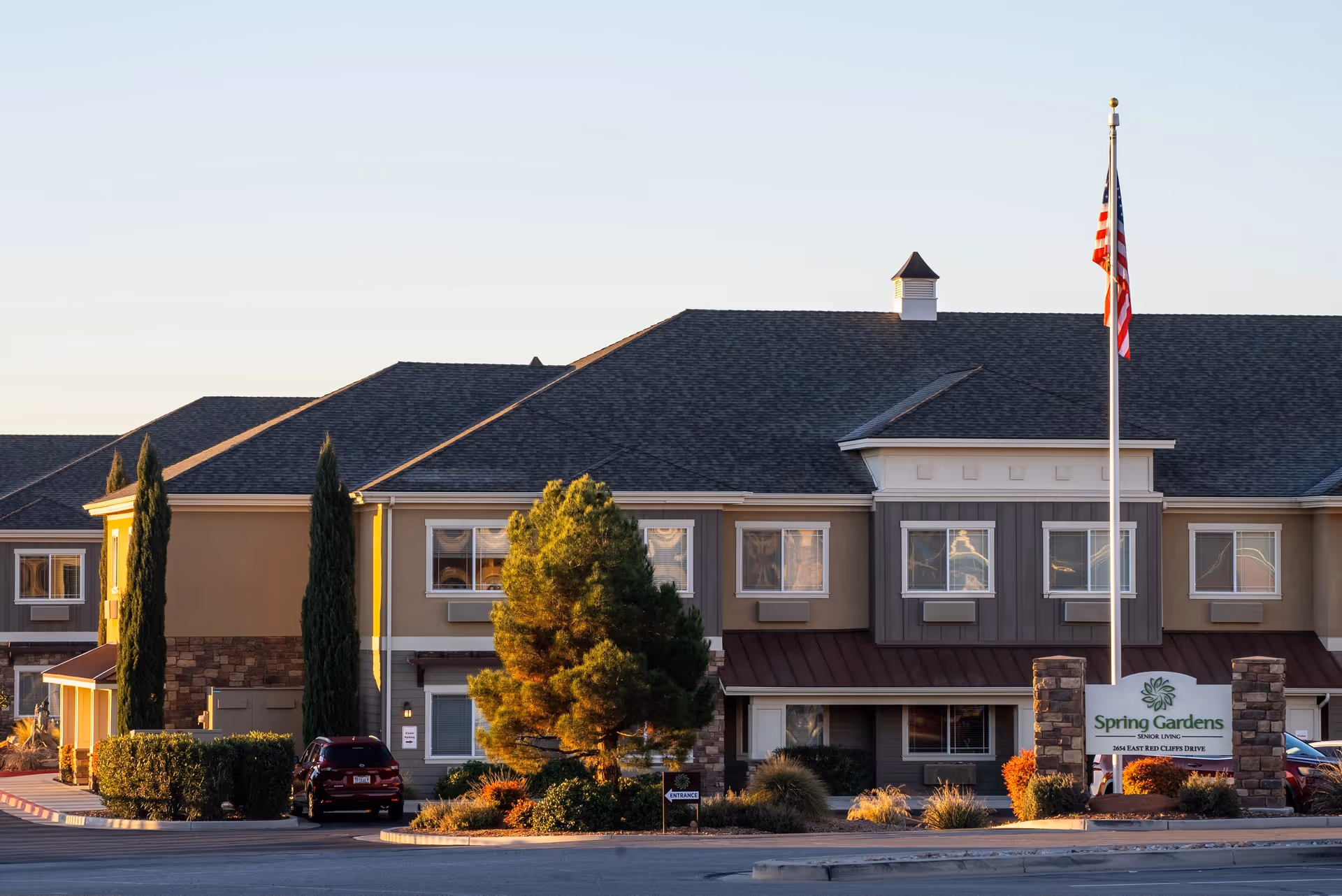Exterior view of Spring Gardens St. George senior living facility showing a two-story building with multiple windows, a landscaped area with trees and bushes, a flagpole with an American flag, and a sign displaying the facility's name and address.