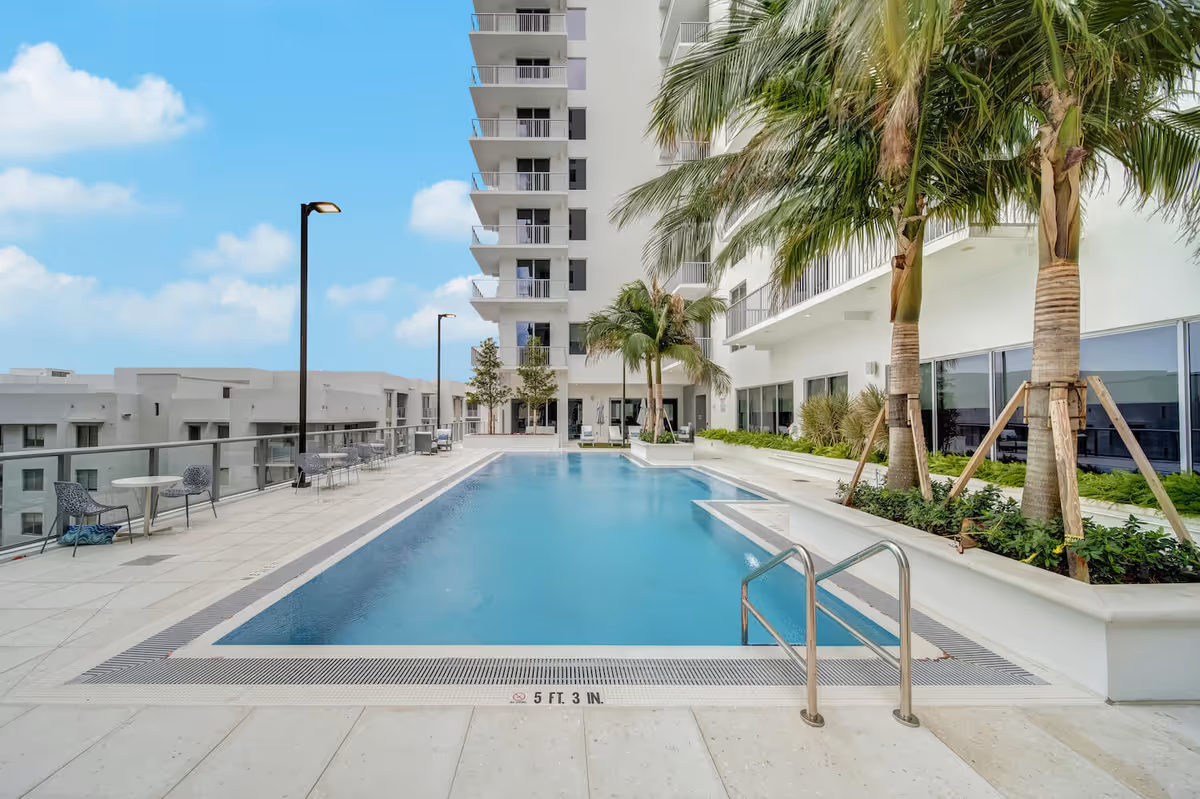 Outdoor swimming pool area with clear blue water, surrounded by a tiled deck with tables and chairs. Tall palm trees and modern white buildings with balconies are visible around the pool under a partly cloudy blue sky.