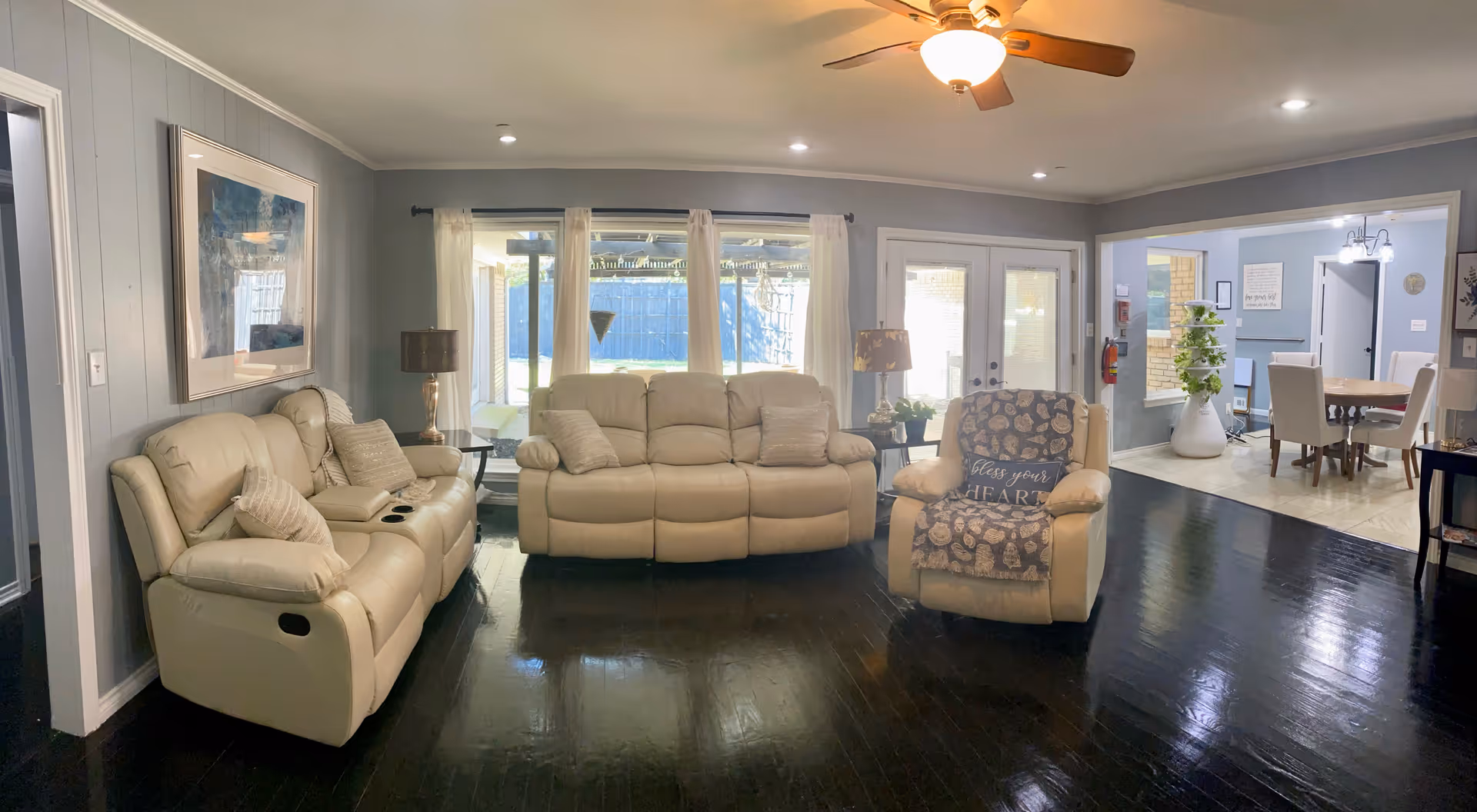A cozy living room with light beige leather recliner sofas and armchair arranged around a dark wooden floor. The room has light gray walls with a large framed picture, two table lamps on side tables, and large windows with sheer white curtains letting in natural light. In the background, there is a dining area with a wooden dining table and chairs.