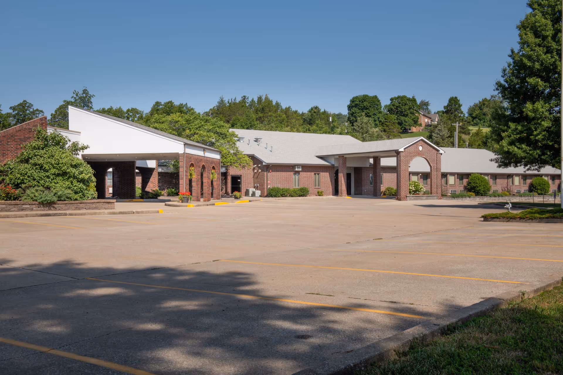 Exterior view of StoneBridge Senior Living - Hermann showing a large brick building with a covered entrance and an empty parking lot in front. The building is surrounded by trees and greenery under a clear blue sky.