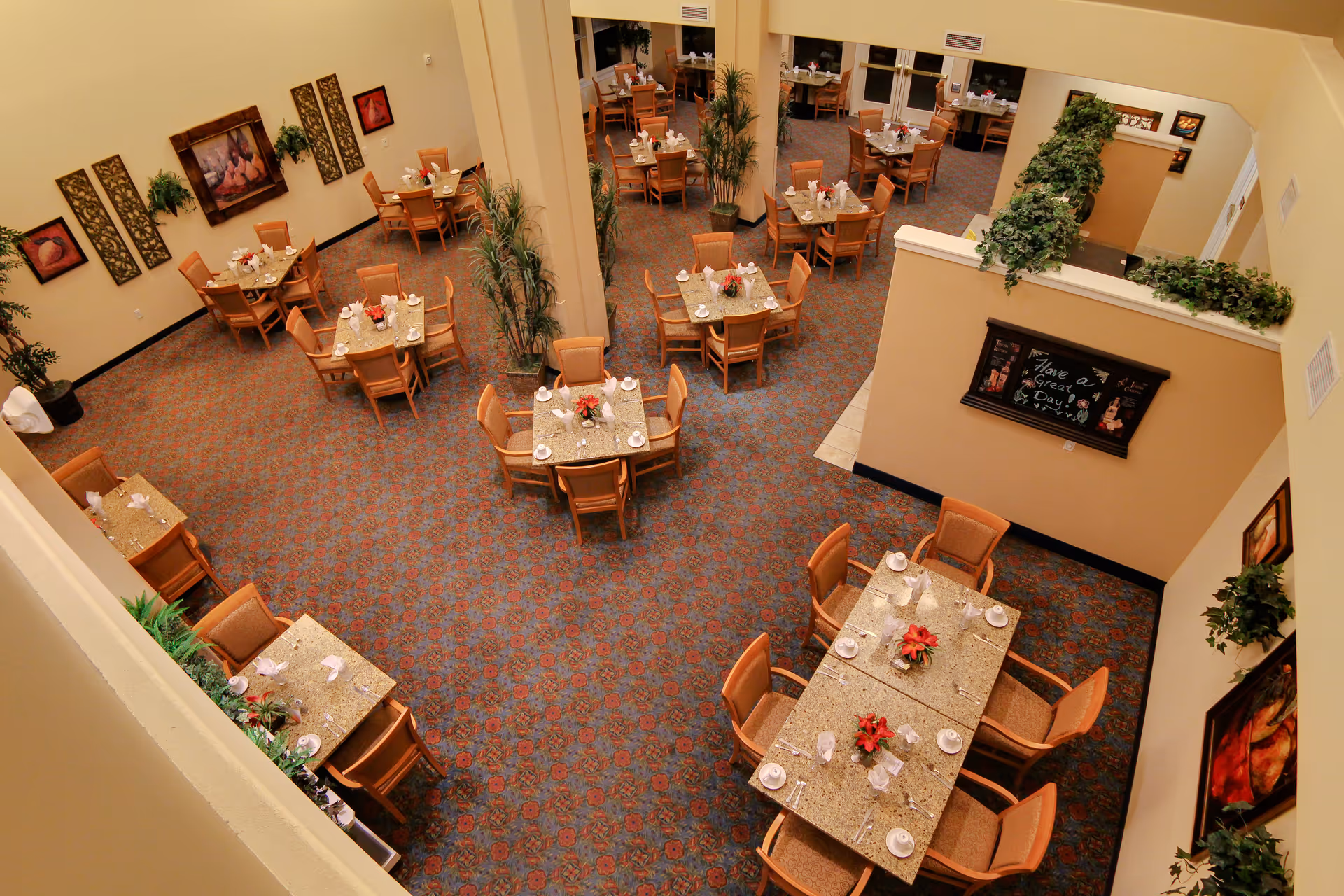 Interior view of a dining room in a senior living facility with multiple tables set for meals, each with chairs, napkins, cups, and small floral centerpieces. The room has patterned carpet, beige walls adorned with framed artwork and plants, and a welcoming atmosphere.
